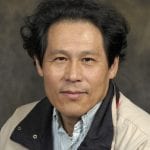 A middle-aged individual with curly black hair, wearing a beige jacket, poses in front of a dark mottled background.