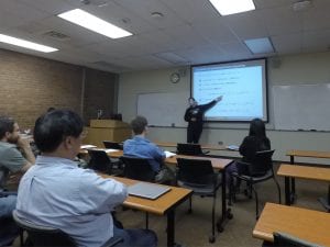 A person stands at the front of a classroom pointing to a large projection screen displaying a presentation. Several people are seated at desks, watching the presentation attentively.