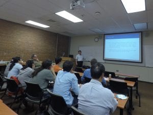 A person is giving a presentation to a group of individuals seated in a classroom. A slide with text is projected on a screen behind the speaker.