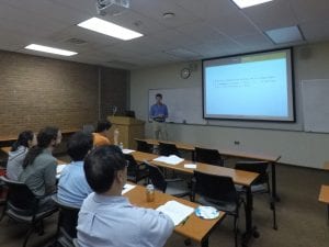 A person is giving a presentation in a small classroom with several attendees seated and looking at a screen displaying text.