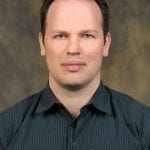 A person with short hair wearing a dark striped shirt against a neutral background.