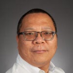 A man with short hair and glasses wearing a white button-up shirt, posed against a plain gray background.