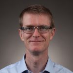 A man with short hair, wearing glasses and a light blue collared shirt, smiles at the camera against a neutral background.