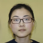 A woman with glasses and dark hair pulled back is facing the camera against a plain yellow background.