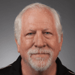 A portrait of an older man with white hair and a beard, wearing a black shirt with a neutral expression against a grey background.
