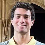 A man with dark curly hair and a light complexion smiles while standing in front of an archway. He is wearing a yellow-collared shirt under a gray sweater.