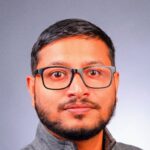 Man with glasses and a beard, wearing a gray zip-up jacket, in front of a gray background.