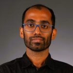 A person with short hair, a beard, and wearing blue glasses and a dark shirt, stands against a gray background.