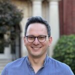 A man wearing glasses and a blue patterned shirt stands outdoors, smiling at the camera with blurred greenery and building columns in the background.