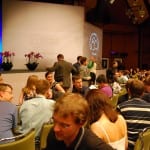 Audience members socializing in a conference hall with a stage displaying a logo in the background.