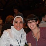 Two women smiling at a table, one wearing a hijab and one with glasses.