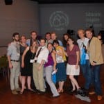 Group of people posing for a photo at a conference, smiling, with a presentation screen in the background.