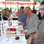 Group of people of various ages sitting at a restaurant table with food and drinks, smiling and enjoying a meal together under an umbrella.