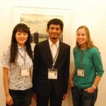 Three people wearing conference badges standing in front of a painting, smiling at the camera.