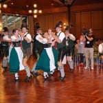 People in traditional bavarian attire dancing at a cultural event in a hall, with onlookers watching and taking photos.