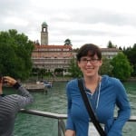 Woman smiling in front of a scenic lake and historical building with a tower, under an overcast sky.