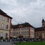 A cloudy day with people gathered at a square surrounded by traditional european-style buildings and a church with a red roof.