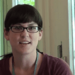 Woman with short dark hair and glasses, wearing a purple top, smiling slightly in front of a window with white curtains.