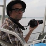 Man in plaid shirt and hat holding a camera on a boat, with a lake in the background.