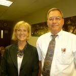 Two smiling middle-aged individuals, a woman and a man, standing side by side in a room with wood-paneled walls.