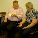 Two people conversing in an office setting, one sitting with a plate of food and the other attentive.