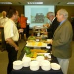 Group of men standing at a buffet table with various food items in a room with a presentation screen displaying an image of a house.