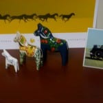 Three decorative wooden horses, each painted differently, displayed in front of a picture with horse silhouettes and a postcard featuring a tractor.