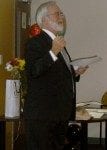 An elderly man with a beard, wearing a black suit and glasses, giving a speech while holding papers in a room with flowers in the background.