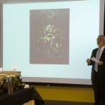 A man stands presenting in a conference room with a projection of a floral image on the screen behind him.