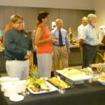 Group of adults standing near a buffet table with food, in a casual event setting.