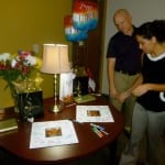 A man and woman examining documents on a table with a lamp, a bouquet of flowers, some balloons, and a small cake in an office setting.