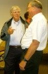 Two elderly men in casual attire engaged in a friendly conversation in a room with beige walls.