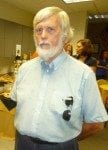 Elderly man with white beard and hair, wearing a light blue shirt and glasses, standing in a room with people in the background.