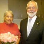 An elderly woman in a red sweater holds a bouquet of pink flowers, standing next to an elderly man in a suit with a pin on his lapel.