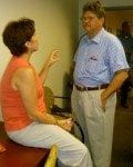 A woman seated on a chair in conversation with a standing man in an office setting.