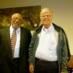 Two elderly men smiling and standing side by side in a room, one in a suit and the other in a casual jacket and glasses.