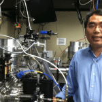 Asian man in a blue shirt smiling in a laboratory filled with intricate scientific equipment.