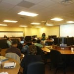 A classroom with students seated at computers, focusing on a presentation given by an instructor at the front.