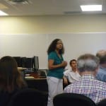 A woman stands presenting to a group of seated adults in a casual, office-like setting.