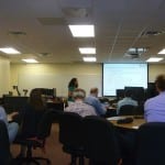 A woman presents to a small audience in a conference room with computers, using a projector screen displaying a graph.