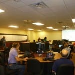 Office training session with multiple participants using laptops and focusing on a presentation at the front of the room.