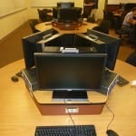 Office workspace featuring dual monitor computer setup on a desk with partition walls, and background showing people working at similar stations.