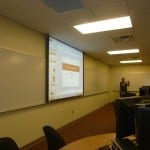 A university classroom with rows of computers and a projector screen displaying a presentation, with a lecturer standing at the front.