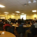 A classroom setting with adults seated at computers, listening to an instructor presenting at the front.