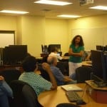 A woman stands presenting at the front of a computer lab with seated adults looking towards her.