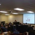 A presenter discussing a bar graph on a projector screen to an attentive audience in a conference room.