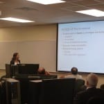 A woman presents a lecture to an audience in a conference room with a powerpoint slide visible in the background.