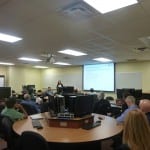 A classroom setting with people seated at tables facing a presenter at the front next to a projection screen.