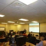 A classroom with adults seated at desks, looking towards a presenter at the front near a projected screen.