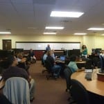 Students and instructors in a computer lab with desks and monitors, focusing on a presentation at the front.